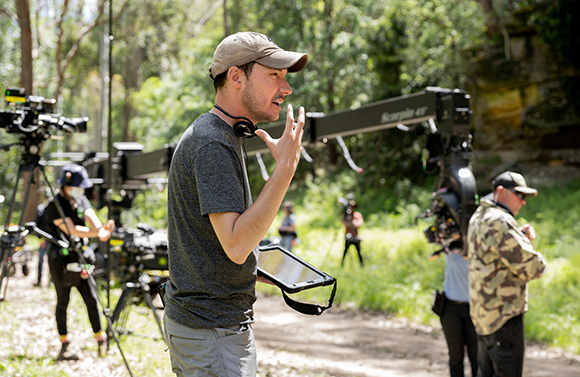 A man in a grey shirt and cap gestures with his hand while speaking on a film set outdoors.