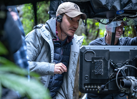 A man in a grey jacket and tan cap wearing headphones looks at a large monitor outdoors.