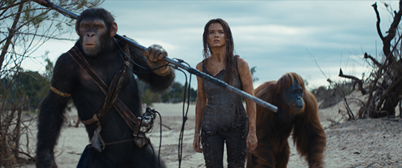 A woman stands between a chimpanzee holding a staff and an orangutan in a sandy, desolate environment.