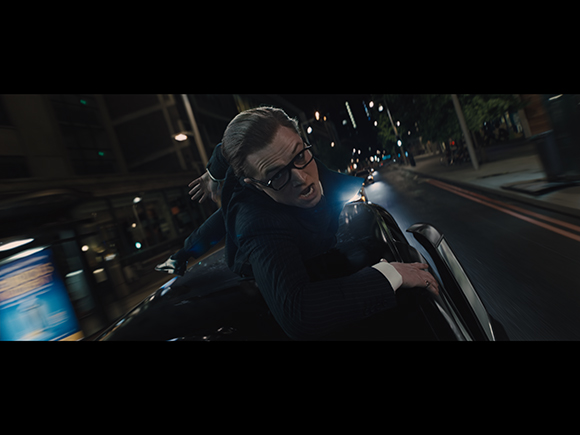 A man with glasses clings to the side of a dark car driving down a city street at night.