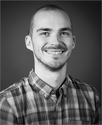 A smiling man with short hair and a beard wearing a plaid button-down shirt against a dark background.