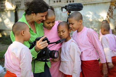 A woman in a green shirt holds a video camera and shows it to a group of young nuns wearing pink and white robes.