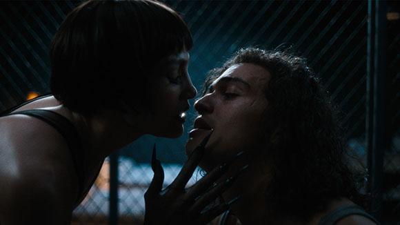 A person with short dark hair leans toward a person with long curly hair in a dark, fenced setting.