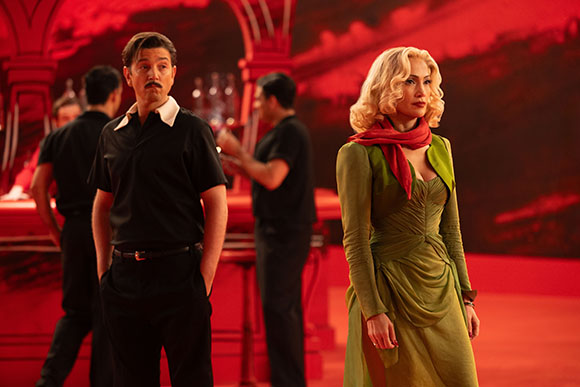 A blonde woman in a green dress stands in a red-lit room near men in black shirts.