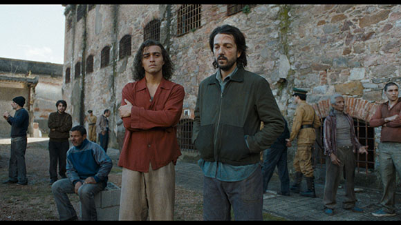 Two men stand in a courtyard before a large stone building with several other men nearby.