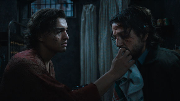 A man in a red robe uses a cloth to wipe the face of a wounded man in a dimly lit room.