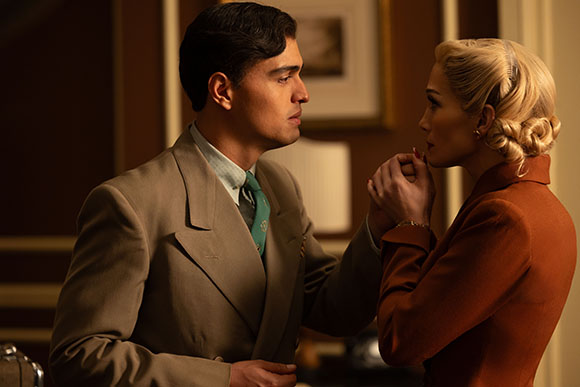 A man in a tan suit and a woman in a rust-colored jacket hold hands and look into each other's eyes.
