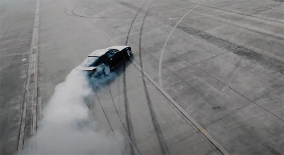 A black car performs a drift on a concrete surface, leaving a thick trail of white smoke.