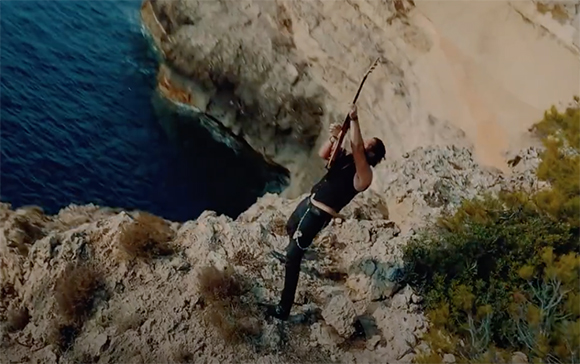 A man in black clothing plays an electric guitar on a rocky cliff overlooking the ocean.
