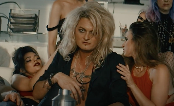 A person with large blonde hair and dark eyeliner sits among several people in a dimly lit setting.