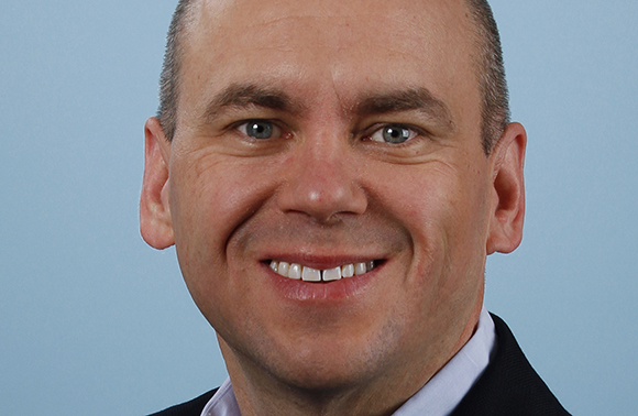 A smiling man with short hair and blue eyes looking toward the camera against a light blue background.
