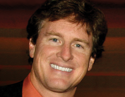 A smiling man with wavy brown hair and blue eyes wearing an orange shirt.