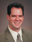 A smiling man with dark hair wearing a tan suit jacket, white shirt, and patterned tie.