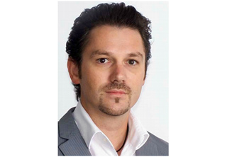 A man with dark wavy hair and a goatee wearing a white shirt and grey suit jacket.