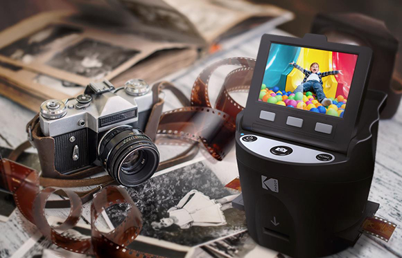 A vintage film camera and loose film strips surround a modern black digital photo scanner displaying a colorful image.