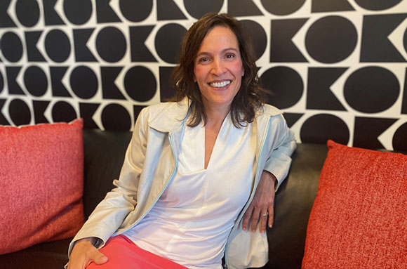 A smiling woman in a white shirt and light jacket sits on a black couch between two red pillows.
