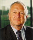 A smiling man with graying hair and a beard wearing a dark suit, light blue shirt, and striped tie.
