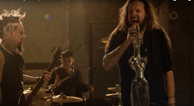 A vocalist sings into a mannequin torso microphone while a guitarist and drummer perform in the background.