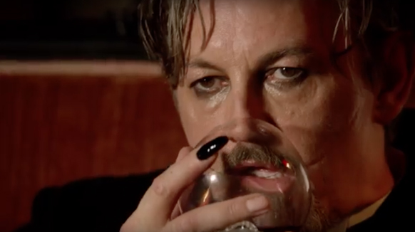 Close-up of a man with light hair and a goatee drinking from a clear glass with black fingernails.