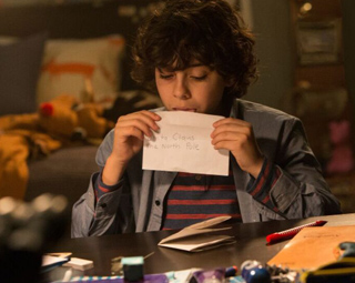 A young boy with curly hair holds a handwritten letter that says 'To Santa Claus, North Pole'.