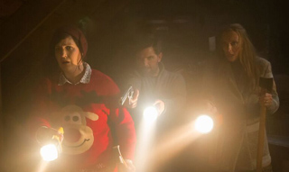 Three people in a dark room hold bright flashlights, looking ahead with concerned expressions.