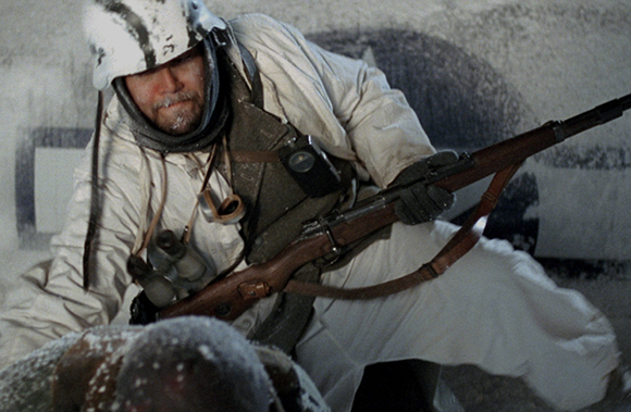 A soldier in white winter gear crouches behind cover while holding a rifle.