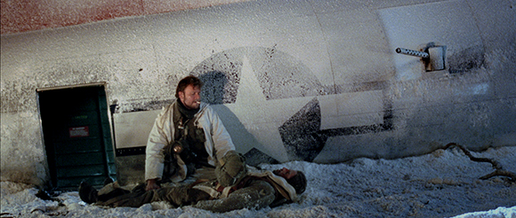 A man in a white jacket sits beside a person lying on the snow next to a large plane fuselage.