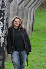 A woman with glasses and curly hair leans against a weathered concrete wall outdoors.