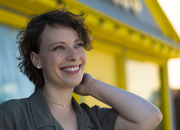 A smiling woman with short curly hair looking up and to the side against a yellow building background.