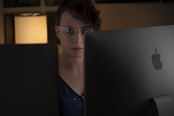 A woman wearing glasses looks intently at a computer monitor in a dark room.