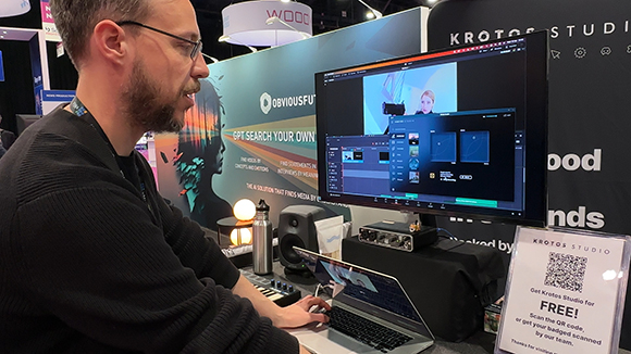 A man in glasses sits at a desk using a laptop and large monitor displaying video software.