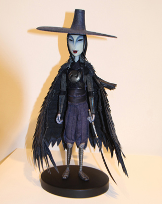 A doll of a character wearing a wide-brimmed hat, a black feathered cape, and dark pants, holding a sword.