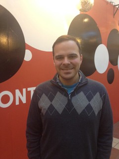 A man wearing a blue quarter-zip sweater with a grey argyle pattern smiles at the camera. He stands in front of a red wall wi