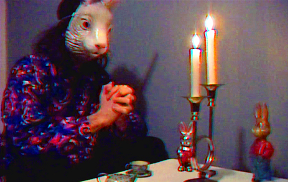 A person wearing a white animal mask and patterned shirt sits near two lit candles on a table.