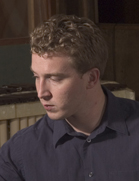A young man with curly light brown hair wearing a dark blue button-down shirt looking downward.
