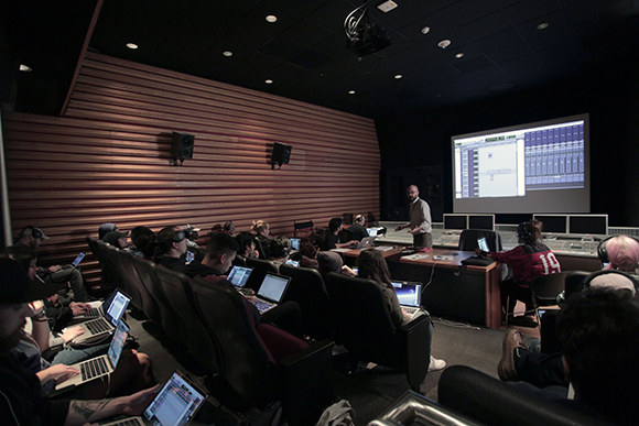 People sitting in dark theater seats using laptops while watching a digital interface on a large projection screen.