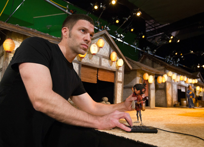 A man in a black t-shirt manipulates a small puppet with his hands on a keyboard in front of a miniature village set with pap