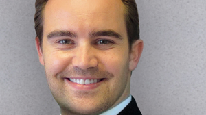 A man with short brown hair and light stubble smiling against a gray background.