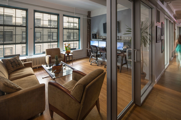 A lounge with tan sofas and armchairs next to a glass-walled office containing desks and computers.