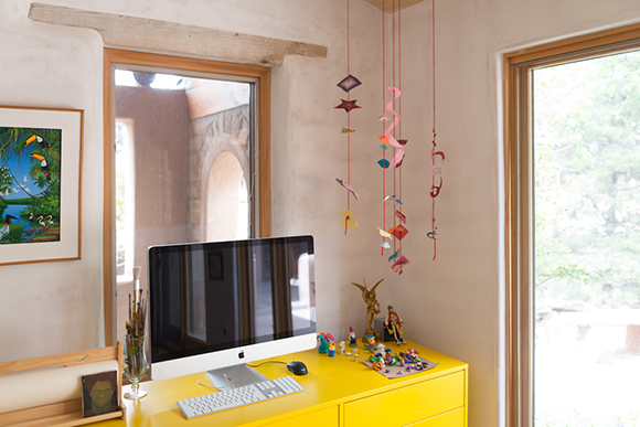 A computer monitor sits on a yellow desk next to a window and colorful hanging decorations.