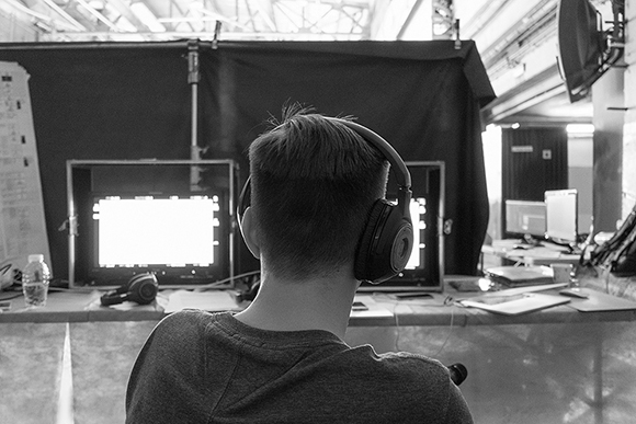 A person from behind wearing headphones, looking at two bright computer monitors in a studio.