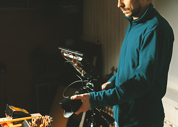 A man in a teal fleece holds a large camera rig with attached monitors and accessories.