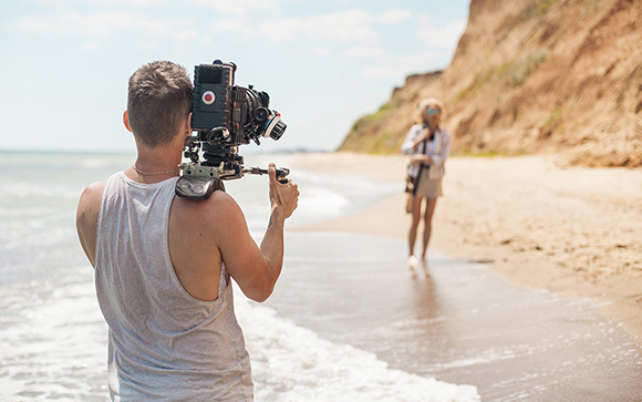 A man in a white tank top holds a professional video camera, filming a woman walking on a sandy beach.