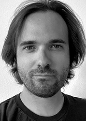 Black and white close-up portrait of a man with dark, shoulder-length hair and a light beard looking at the camera.
