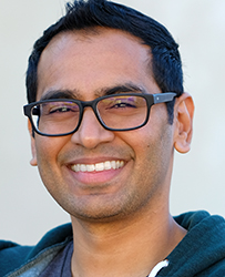 A smiling man with dark hair and black-rimmed glasses looking toward the camera.
