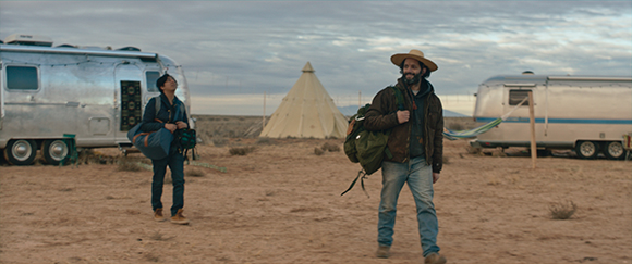 Two people walk across a desert landscape near silver trailers and a large tipi under a cloudy sky.