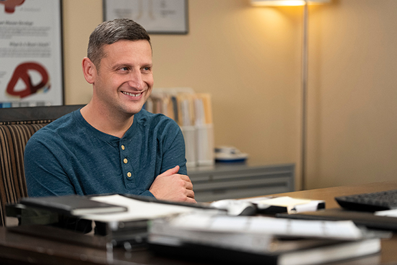 A man in a blue shirt smiles while sitting at a desk in an office setting.