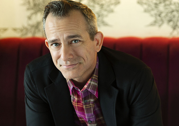 A man with short brown hair wearing a black blazer and a purple plaid shirt smiles at the camera.