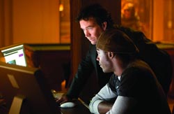Two men lean in to look at a glowing computer screen in a dark room.