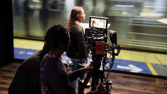 A person with dreadlocks operates a professional video camera to film a woman standing before a large screen.
